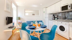 A modern kitchen with white cabinets, a built-in microwave, and a washing machine. There is a dining table with blue chairs and a small side table with a lamp. - Charming Flat in Alfama (Lisbon)