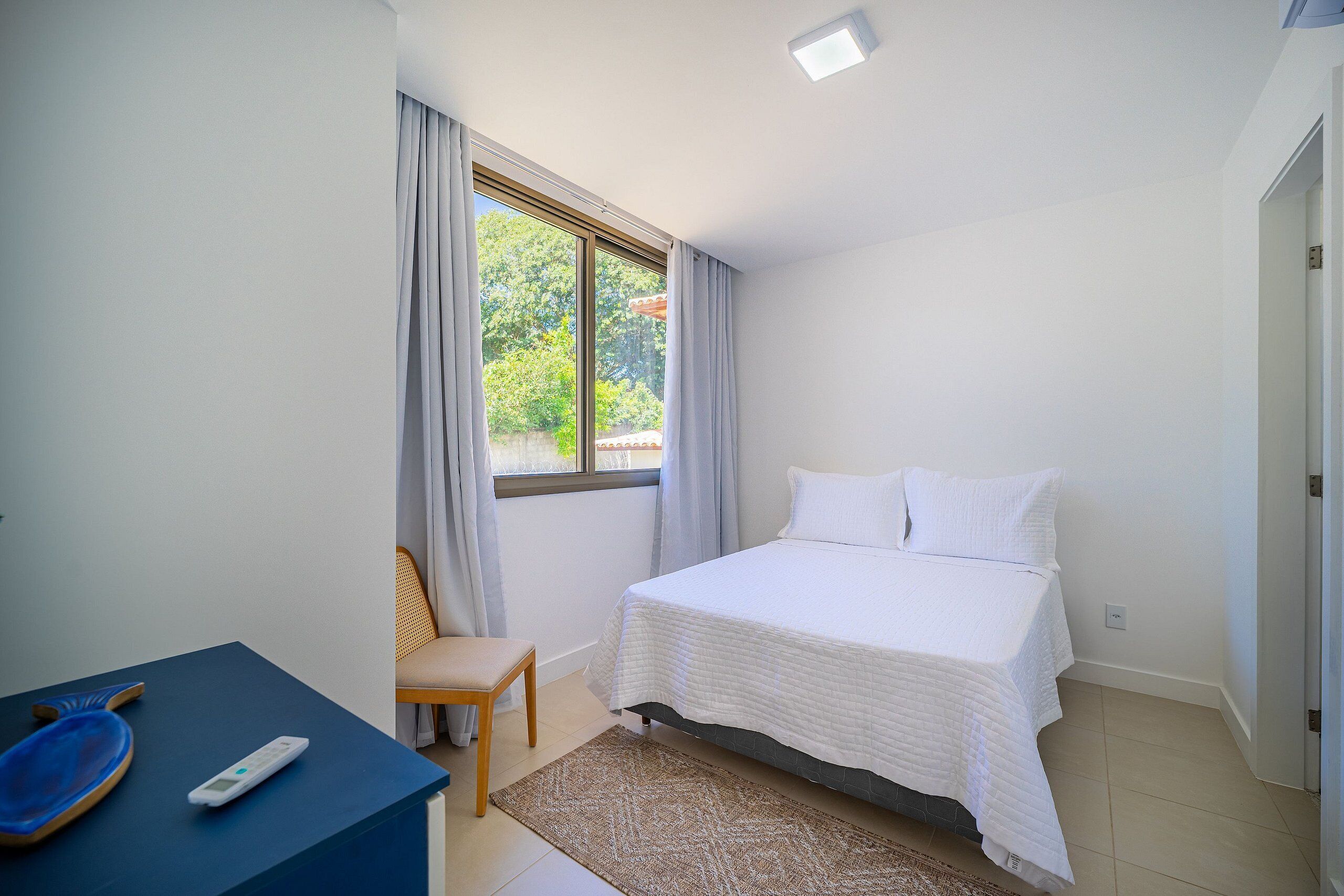 A bedroom with a bed, a chair, a table with a remote control, and a window with curtains.