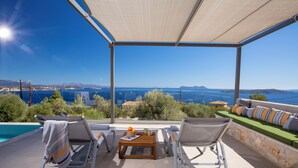 Un área de estar al aire libre con techo, vista a la piscina, sillones de descanso y una mesa con una cesta y vasos.