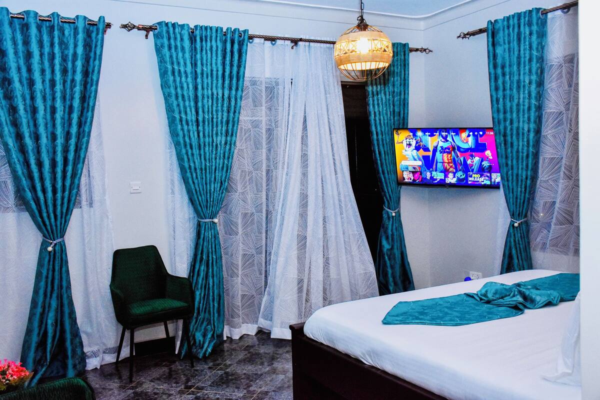 A bedroom with teal curtains, a bed, a chair, and a TV on the wall.