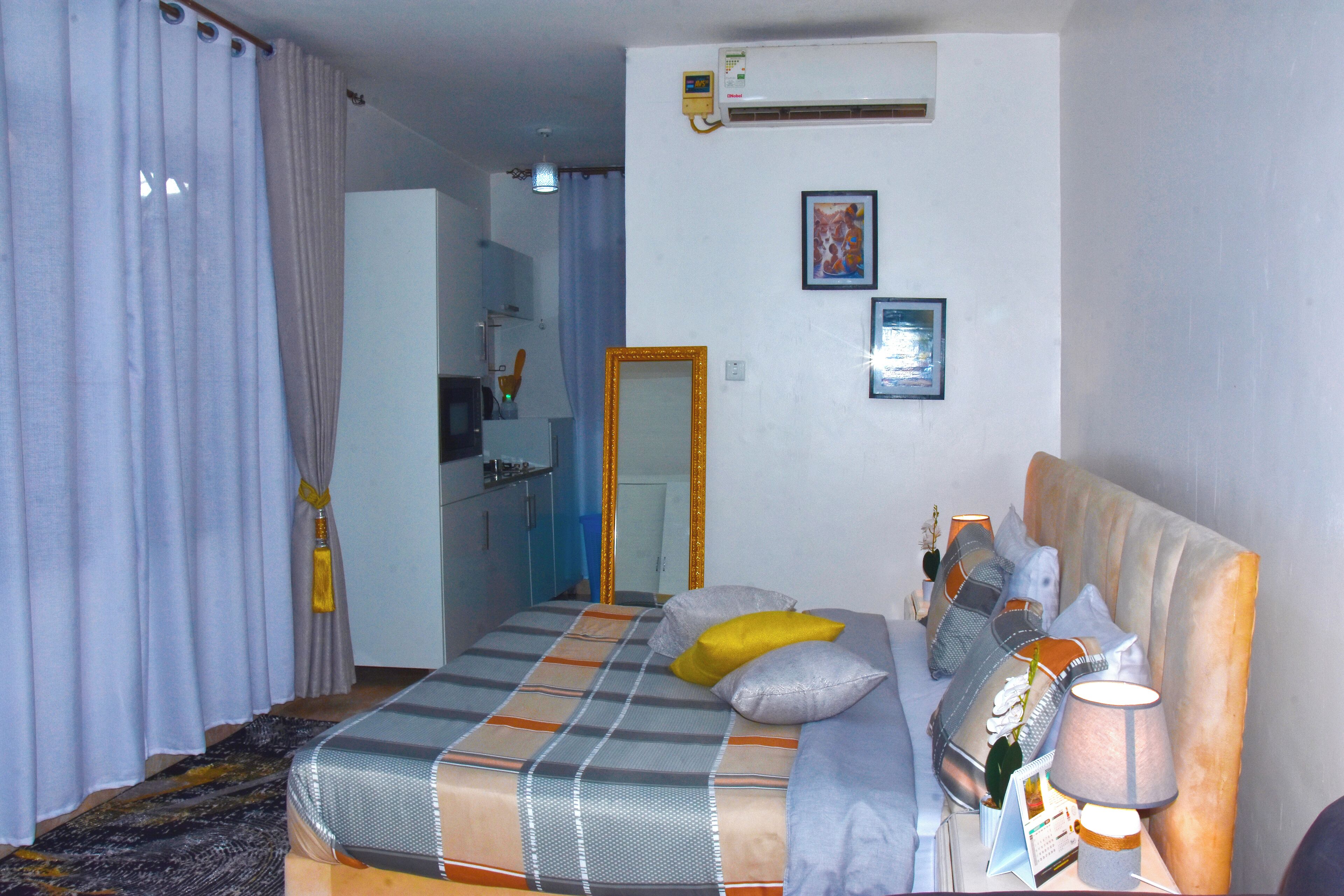 A bedroom with a bed, pillows, a lamp, a mirror, and a wall-mounted air conditioner.