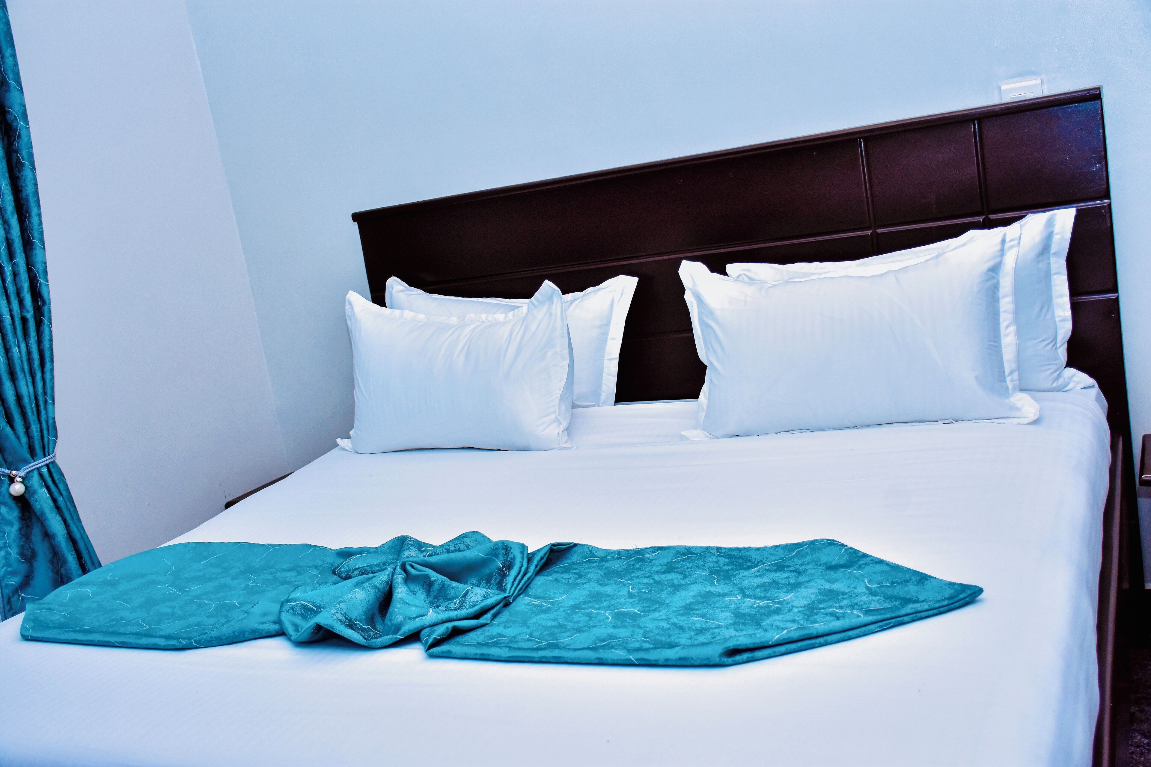 Una cama bien tendida con sábanas blancas y una manta azul turquesa, un cabecero oscuro y una cortina a la izquierda.