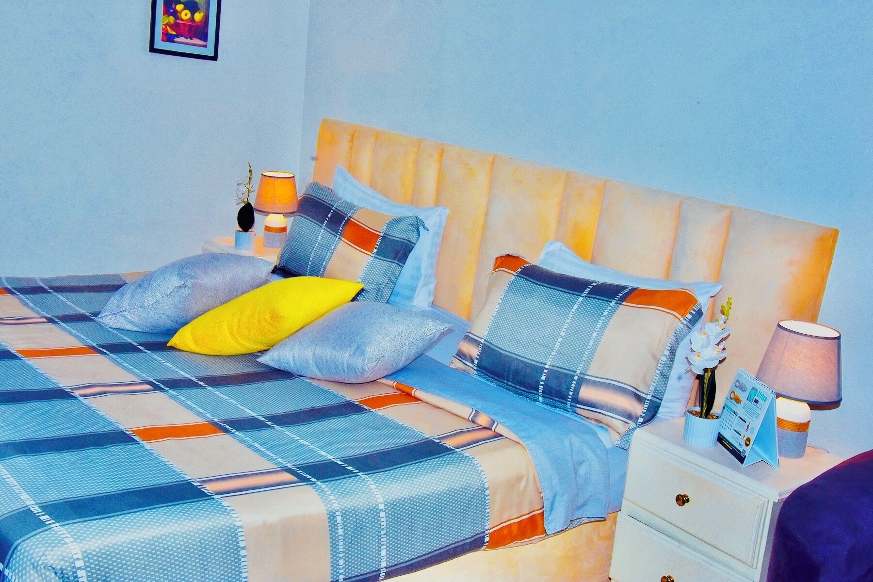 A bed with a blue and orange checkered bedspread, multiple pillows, and a wooden headboard. There is a nightstand with a lamp and a small potted plant.