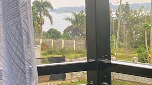 A view from a window with a metal railing, overlooking a body of water, palm trees, and a distant shoreline. - Good deeds Stay (Entebbe)
