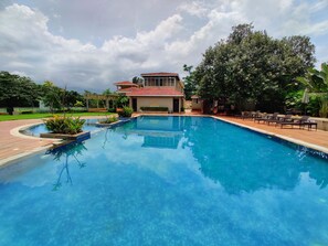A swimming pool with lounge chairs and a building in the background. - Pushpam sanskruti Resort (Karjat)