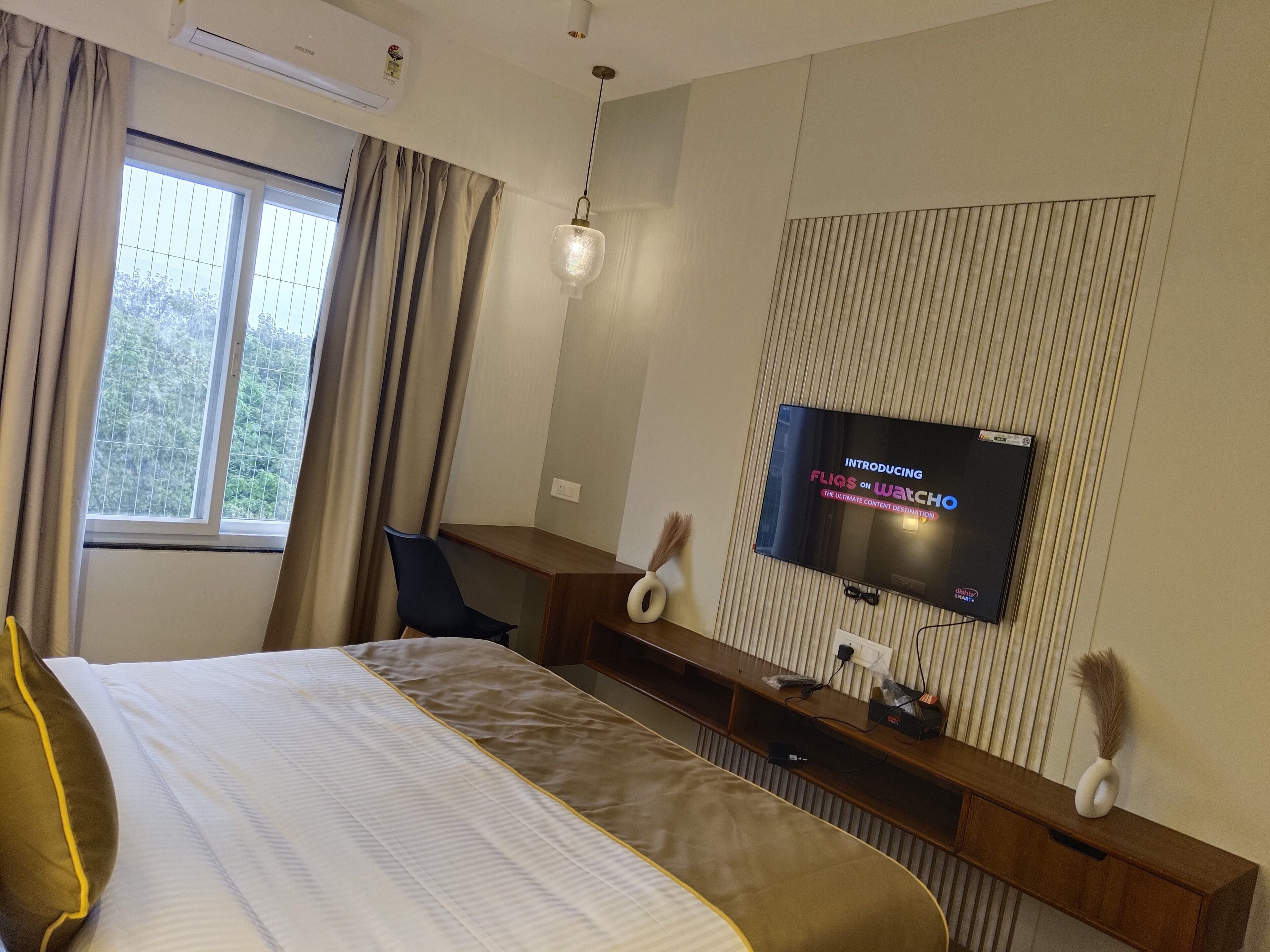 A modern hotel room with a large bed, a desk, a television displaying 'FLIQS ON WATCH', and a window with curtains.