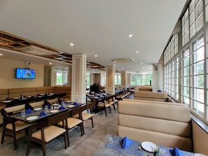 A dining area with long tables, cushioned seating, and large windows. - Pushpam sanskruti Resort (Karjat)