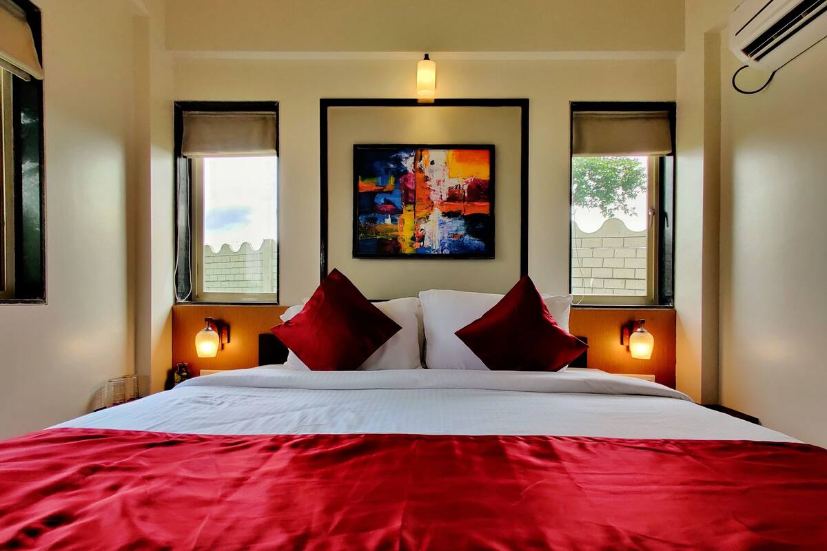 Una habitación de hotel con una cama grande, ropa de cama roja y blanca, dos lámparas de noche, un cuadro sobre la cama y ventanas con cortinas.