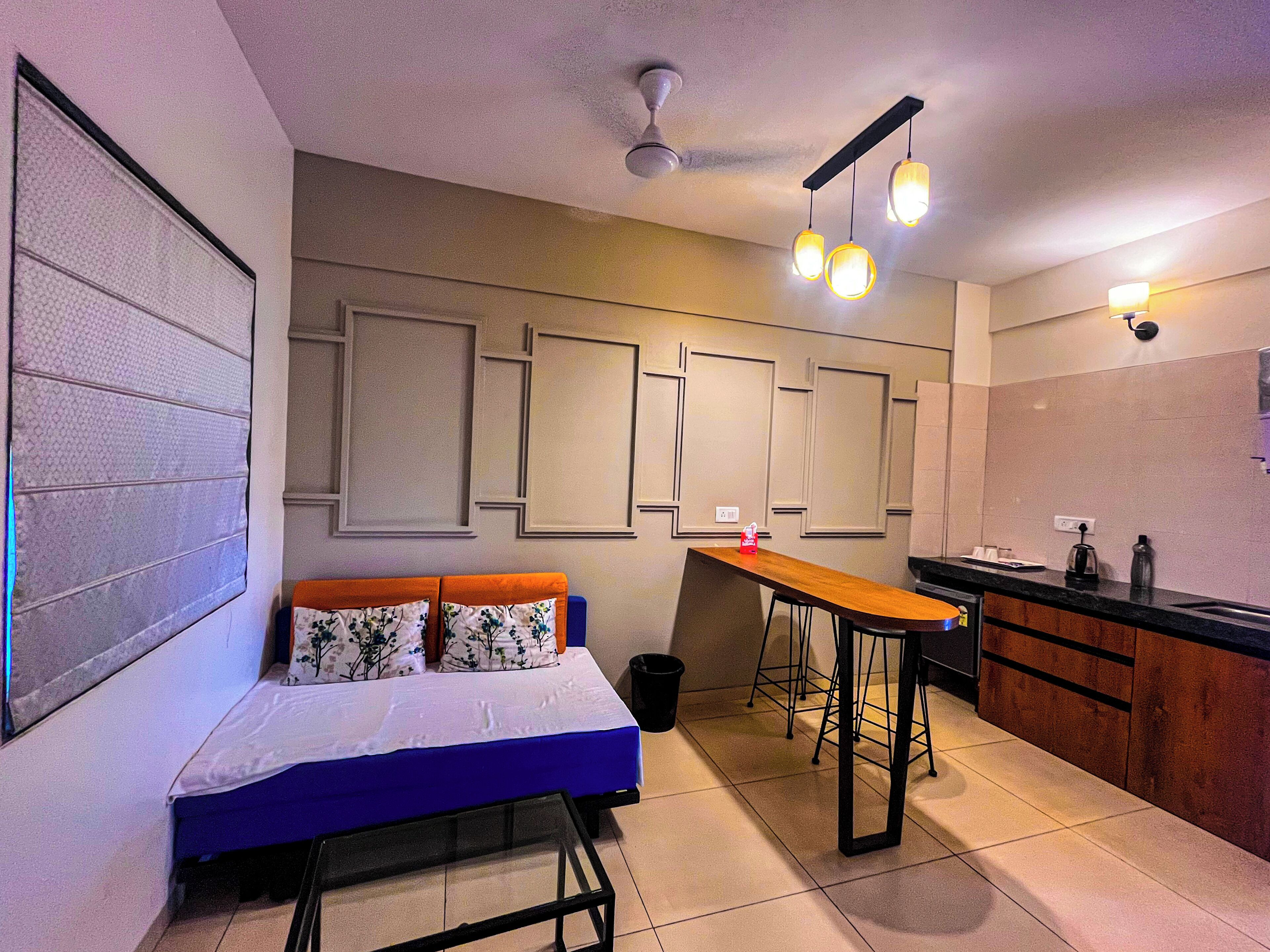 A compact room with a bed, a small table, a kitchenette, and a ceiling fan.