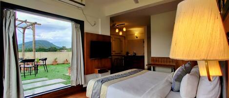 Quarto de hotel com cama, televisão, cabeceira de madeira, varanda com mesa e cadeiras e unidade de ar condicionado.