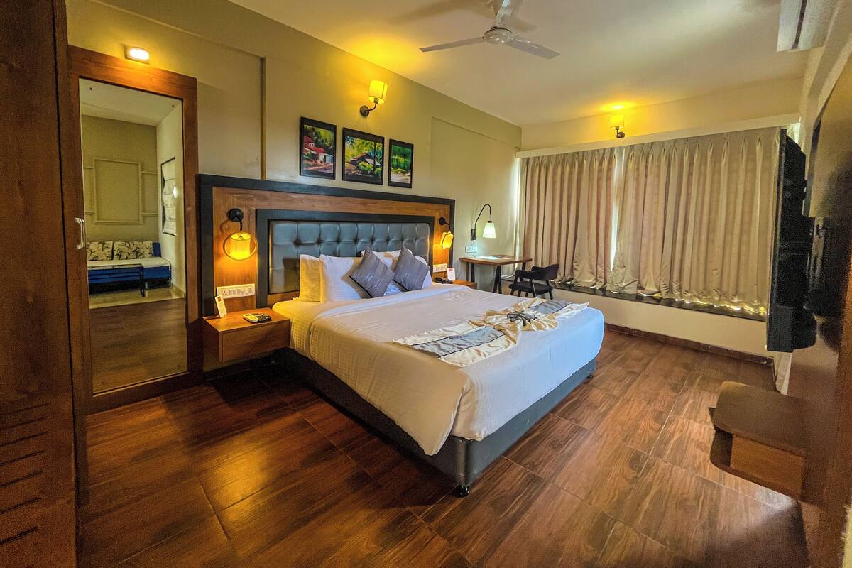 Habitación de hotel con una cama grande, suelo de madera, ventilador de techo y ventana con cortinas.