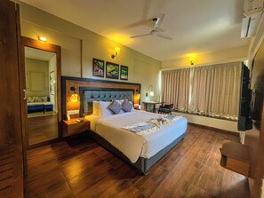 A hotel room with a large bed, wooden flooring, a ceiling fan, and a window with curtains. - Pushpam sanskruti Resort (Karjat)
