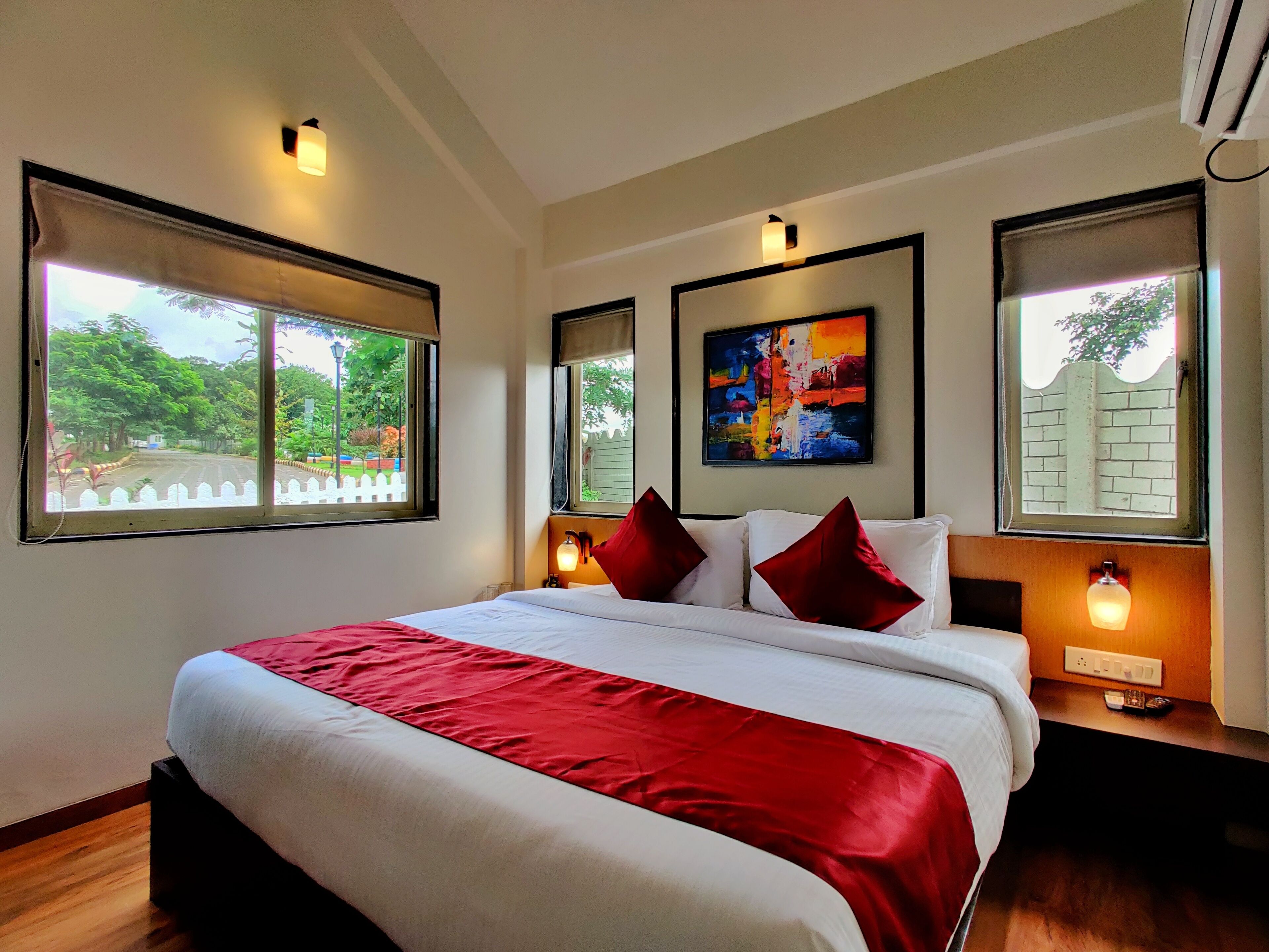 A hotel room with a large bed, two bedside tables with lamps, a colorful abstract painting on the wall, and large windows with a view of greenery.