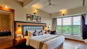 A spacious bedroom with a large bed, a ceiling fan, and a view of a green landscape through the window. - Pushpam sanskruti Resort (Karjat)