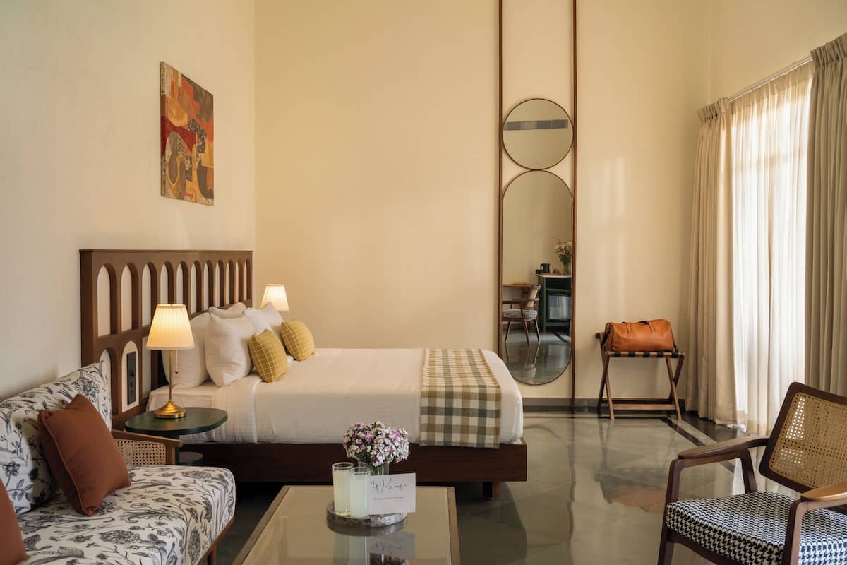 Habitación de hotel con cama, sofá, sillón, mesa de centro, lámpara, espejo y un cuadro en la pared.