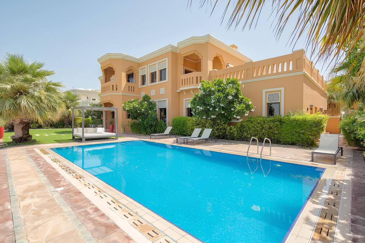 A swimming pool with a tiled deck, a covered seating area, and a building with balconies and a garden.