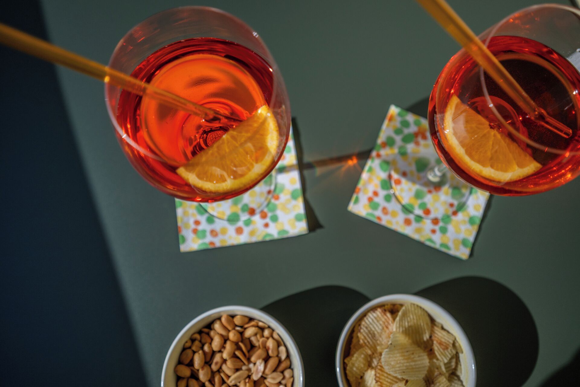 Two glasses of red beverage with orange slices, two bowls of snacks, and colorful napkins on a table.