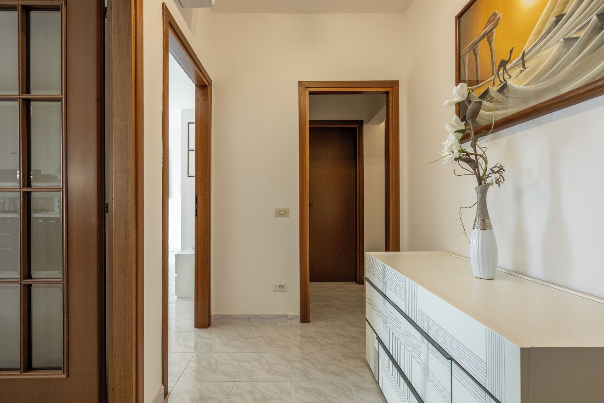 Un pasillo con puerta de madera, un jarrón blanco con flores y suelo de baldosas.