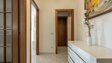 Un pasillo con puerta de madera, un jarrón blanco con flores y suelo de baldosas.