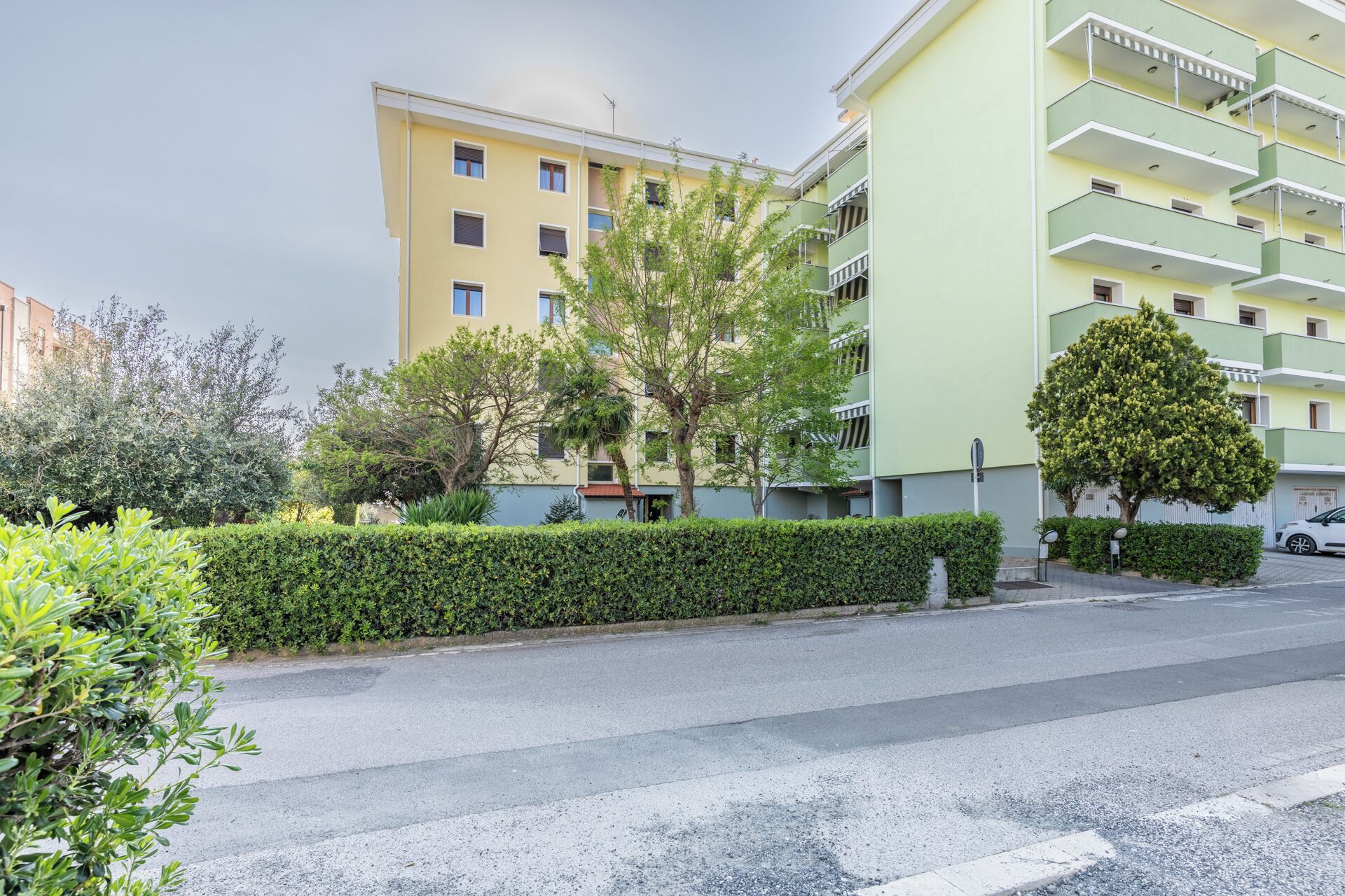 Un edificio residenziale a più piani con balconi, cielo sereno e un cespuglio ben curato lungo la strada.