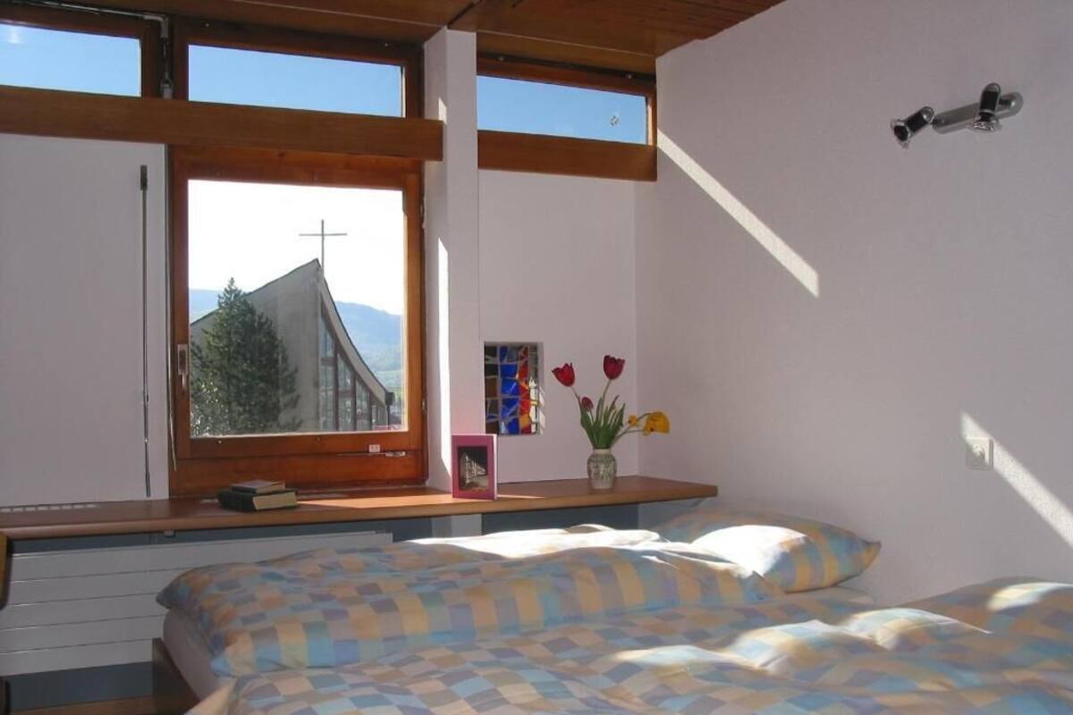 A bedroom with two beds, a window with a view of a mountain and a church, and a small table with a vase of flowers.