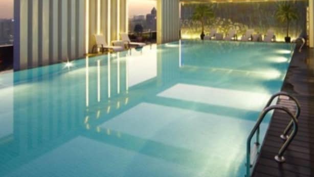 Ein moderner, überdachter Swimmingpool mit klarem Wasser, eine Pool-Lounge mit Liegestühlen und eine Stadtkulisse im Hintergrund.