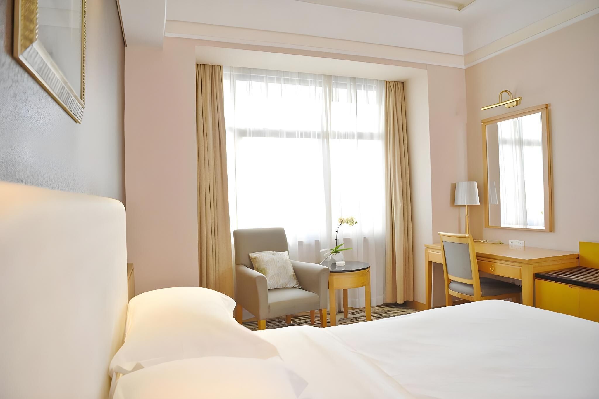 A hotel room with a large bed, a desk with a chair, a small table with a lamp, and a window with curtains.