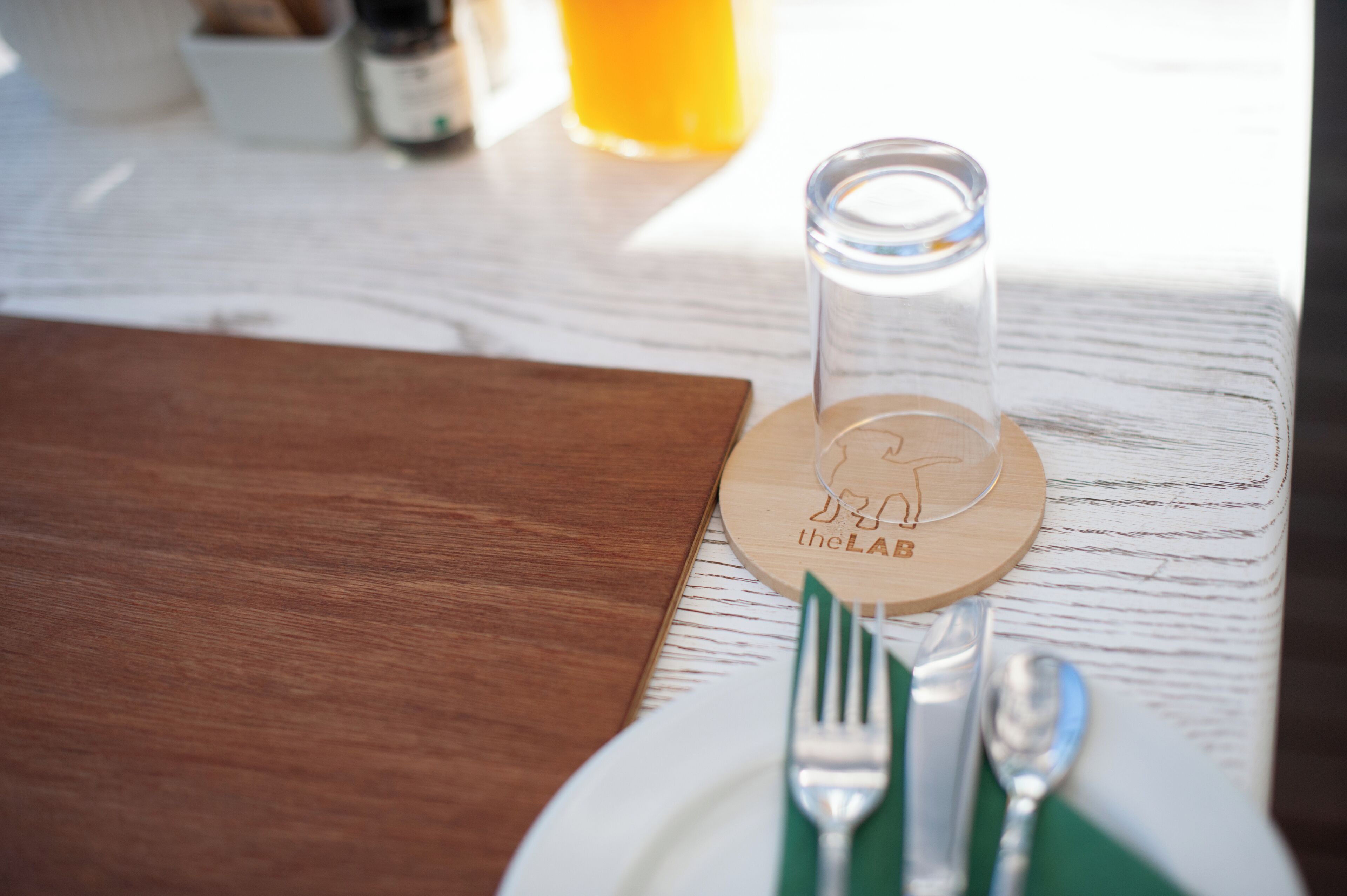 Ein Holztisch mit einem weißen Teller, einer grünen Serviete, einer Gabel und einem Löffel. Außerdem ist ein klares Glas mit Untersetzer zu sehen, das einen Bären und den Text "the LAB" zeigt.