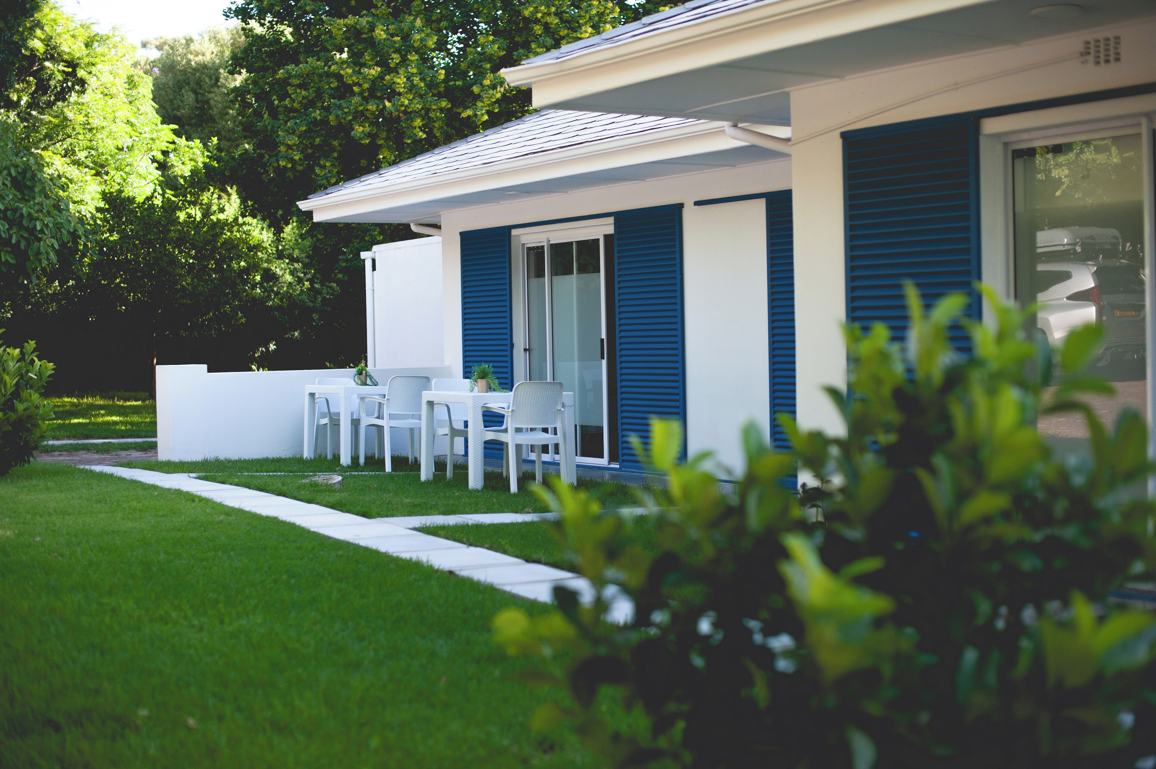 Una casa moderna con fachada blanca y contraventanas azules, un pequeño patio con mesa y sillas, y un césped bien cuidado.