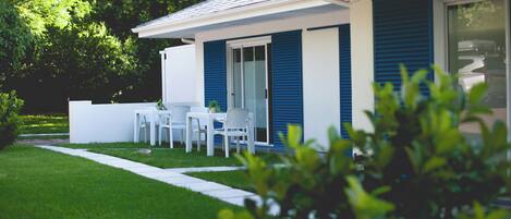 Una casa moderna con fachada blanca y contraventanas azules, un pequeño patio con mesa y sillas, y un césped bien cuidado.