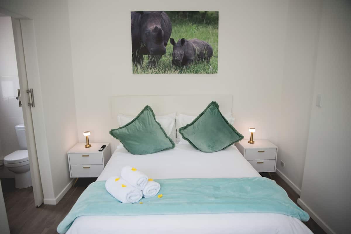 A hotel room with a bed, two bedside tables, a nightstand, a painting of rhinos, and a bathroom visible through an open door.