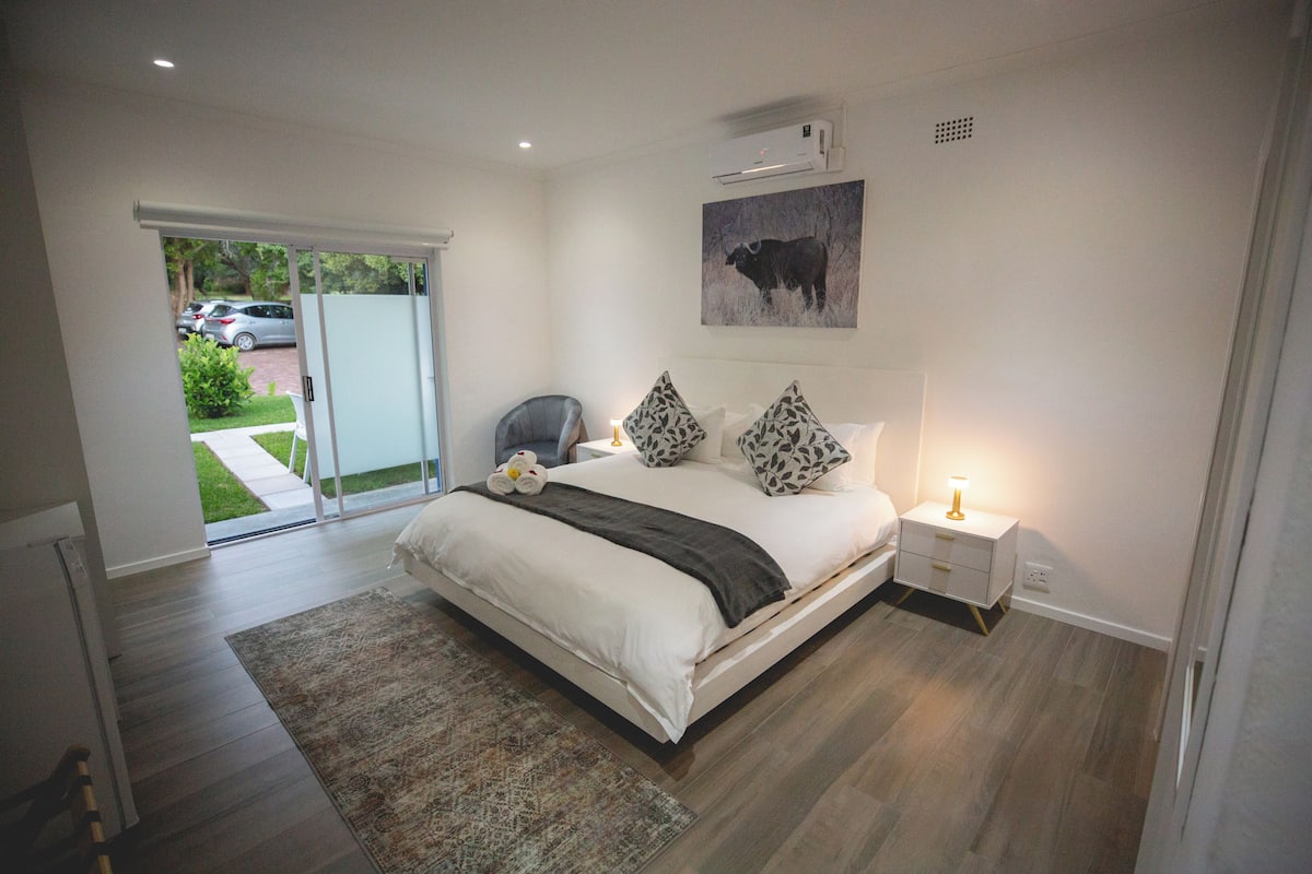 A bedroom with a large bed, a nightstand, a lamp, a wall-mounted air conditioner, and a sliding glass door leading to a patio.