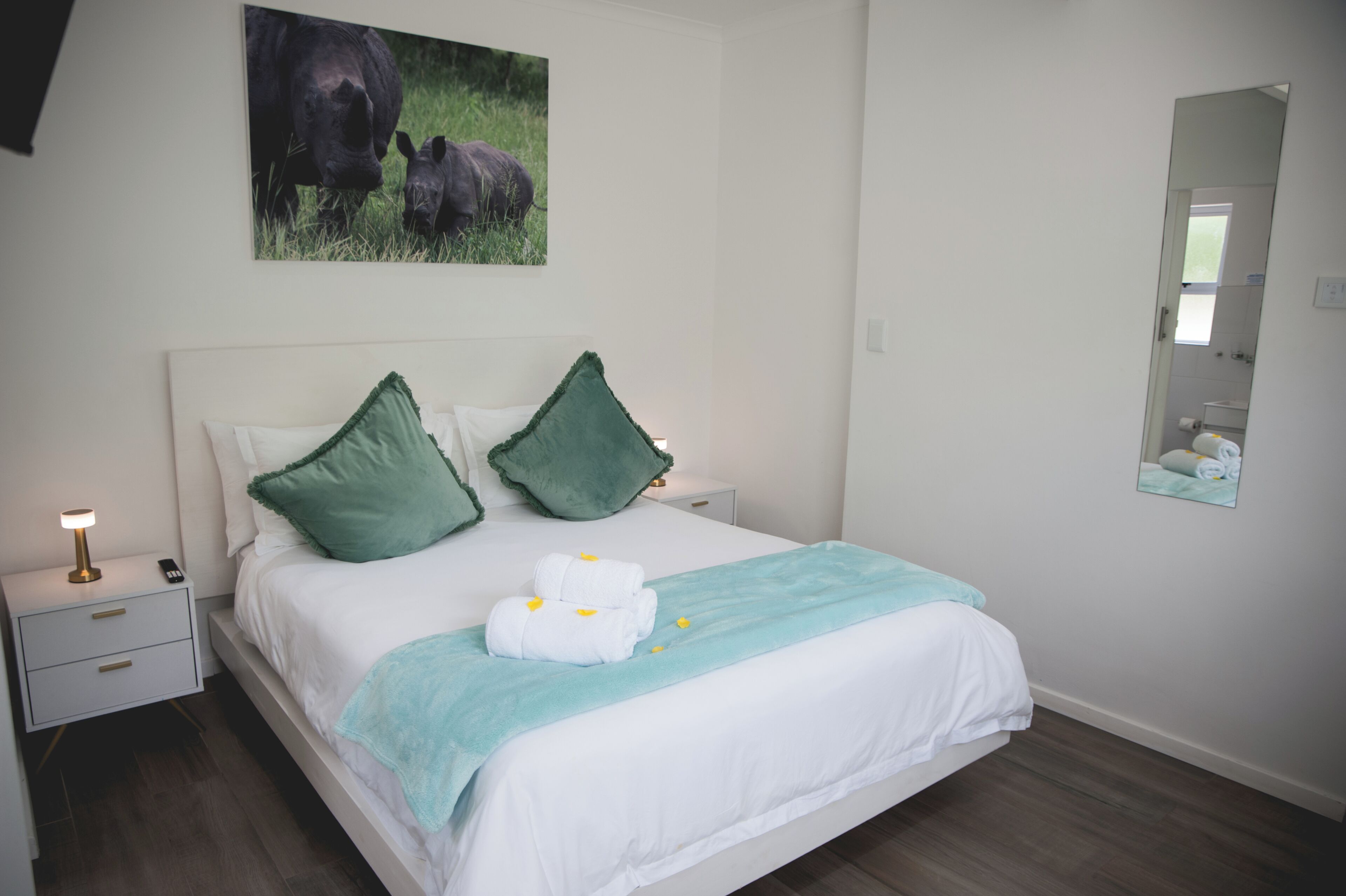 A neatly made bed with white and green bedding, a bedside lamp, and a framed picture of rhinos on the wall.