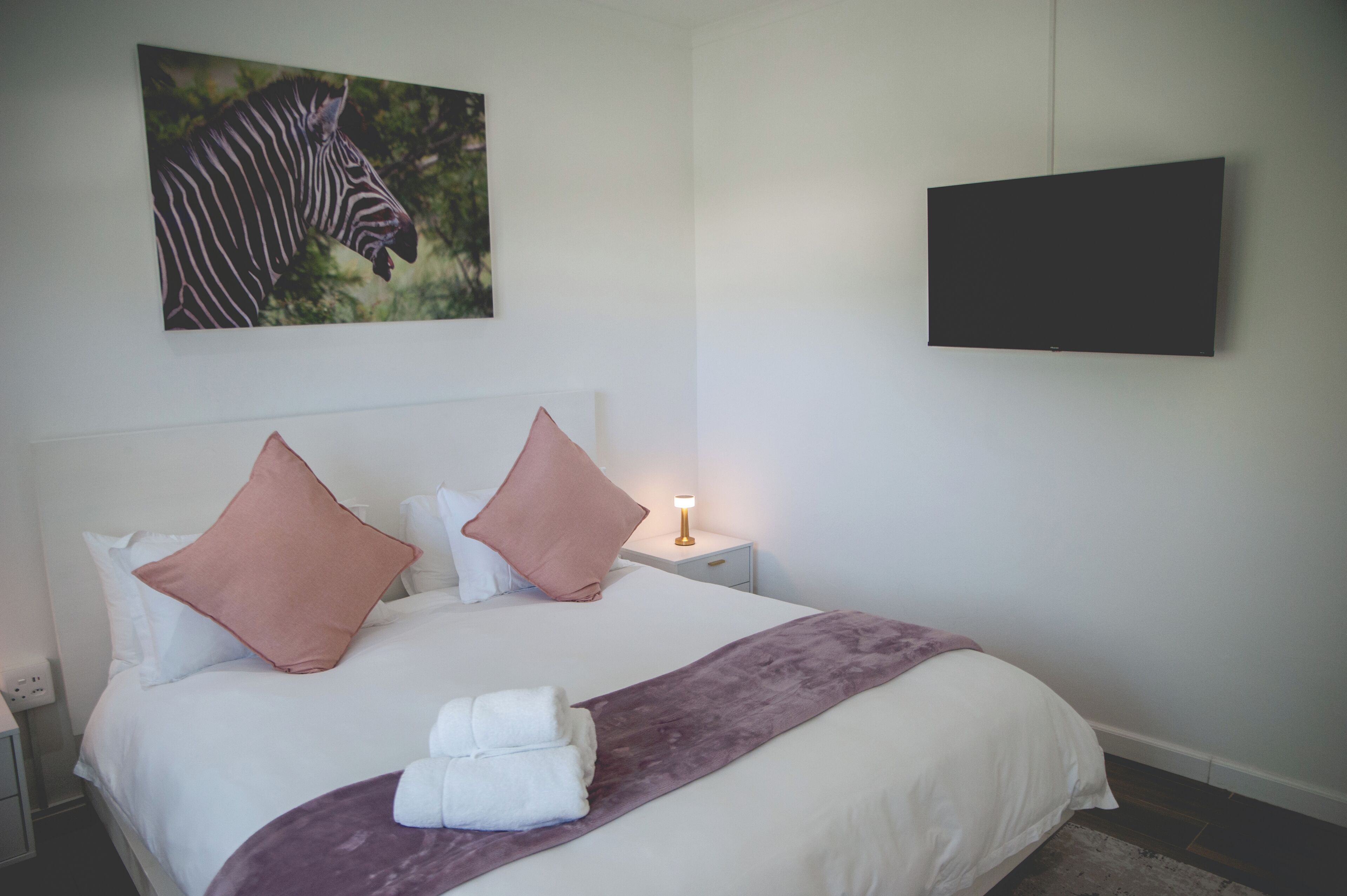 A neatly made bed with white and pink pillows, a purple throw blanket, and a zebra artwork on the wall.
