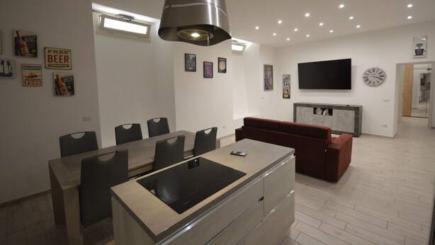 A modern kitchen with a central island, dining area with chairs, a red sofa, and a flat-screen TV mounted on the wall. - SanRemo Apartment (Roma)