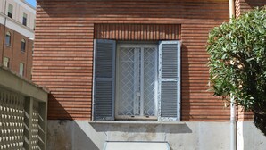 A building with a red brick facade, a window with metal shutters, and a small garden area with a tree. - SanRemo Apartment (Roma)