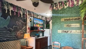 Un salon confortable avec un canapé, une petite table et un fauteuil. Il y a également un coin cuisine équipé d’un réfrigérateur et d’un four à micro-ondes. Les murs sont décorés de motifs floraux et d’une pancarte de bienvenue.