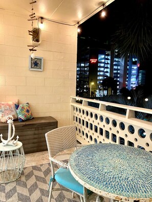 An outdoor seating area with wicker chairs and a round table, a white wall with a framed picture, and a view of a cityscape at night.