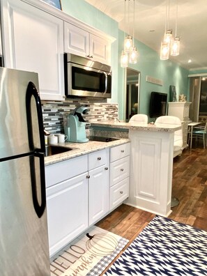 A modern kitchen with white cabinets, a stainless steel refrigerator, a microwave, and a sink. There are hanging pendant lights, a patterned rug, and a door mat with a pumpkin design.