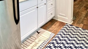 A modern kitchen with white cabinets, a stainless steel refrigerator, a microwave, and a sink. There are hanging pendant lights, a patterned rug, and a door mat with a pumpkin design.