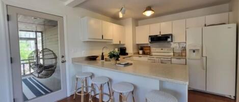 A modern kitchen with white cabinets, a central island with stools, a hanging chair, and a refrigerator.