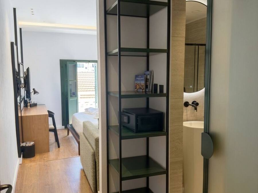 Un intérieur moderne avec un plancher en bois, une étagère en métal noir, un lit, un bureau avec une chaise et une salle de bain avec un miroir et un lavabo.