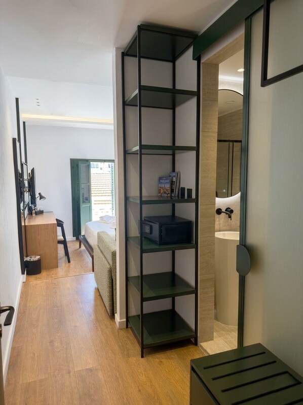 A modern interior with a wooden floor, a black metal shelf unit, a bed, a desk with a chair, and a bathroom with a mirror and sink. - Kentrikon 1924 Boutique Hotel Old Town (Paphos)