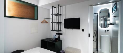 A modern hotel room with a bed, a desk, a TV, a mirror, and a bathroom with a sink and shower.
