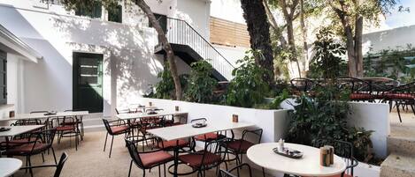 An outdoor cafe with tables and chairs, surrounded by trees and a white building with a staircase.