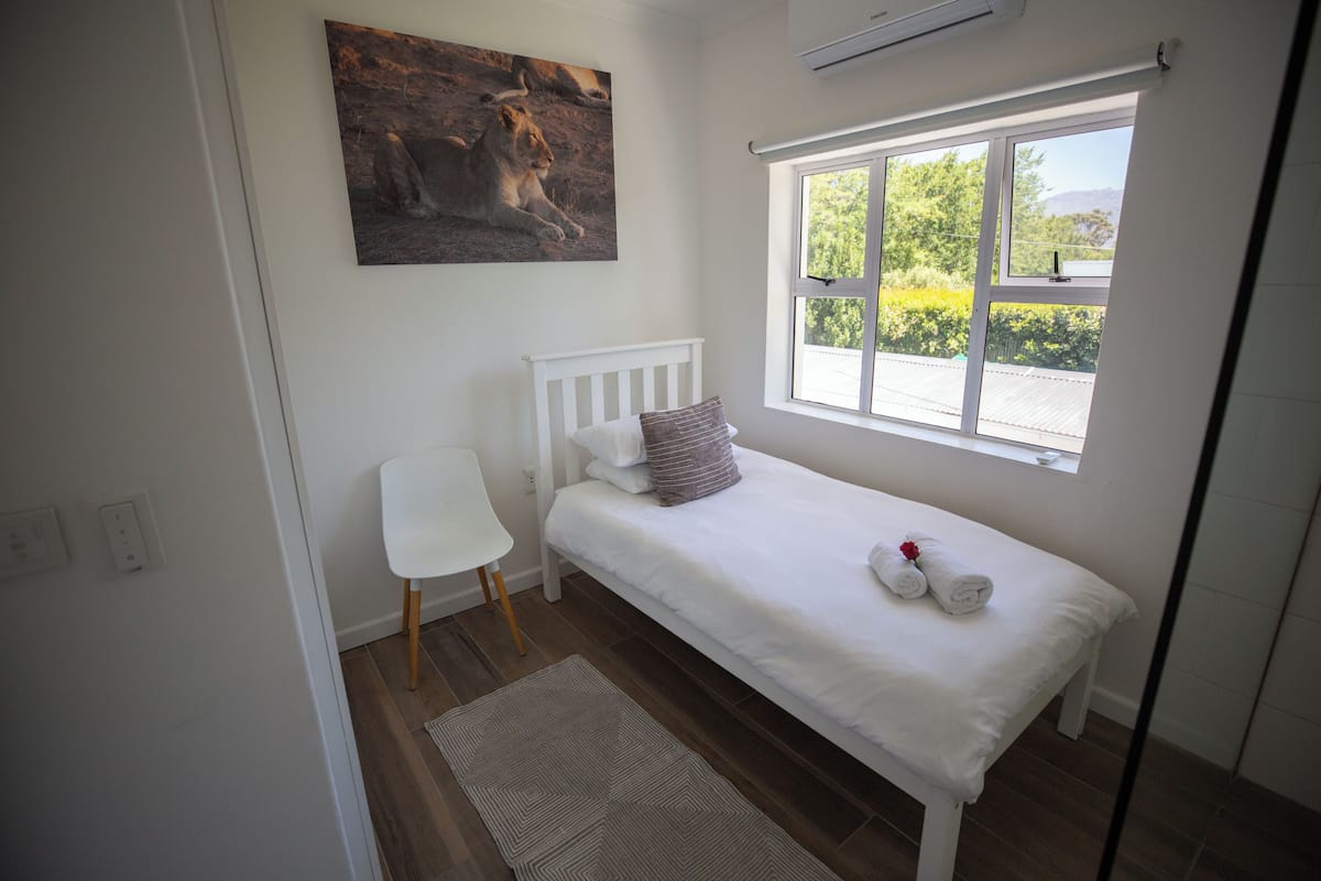 A small, neatly arranged bedroom with a single bed, a window with a view of greenery, a white chair, and a painting on the wall.