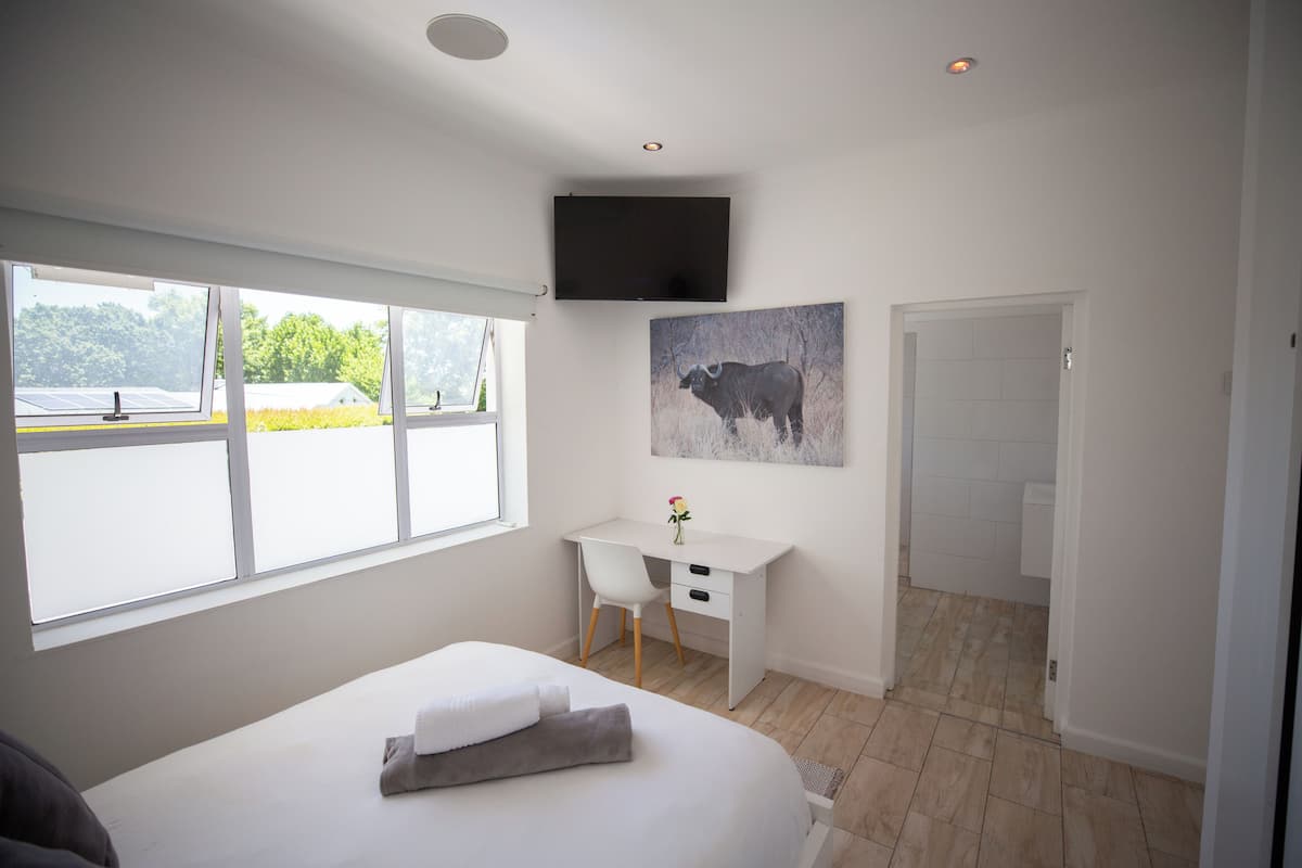 A bedroom with a bed, a desk, a chair, a TV, and a window with a view of greenery.