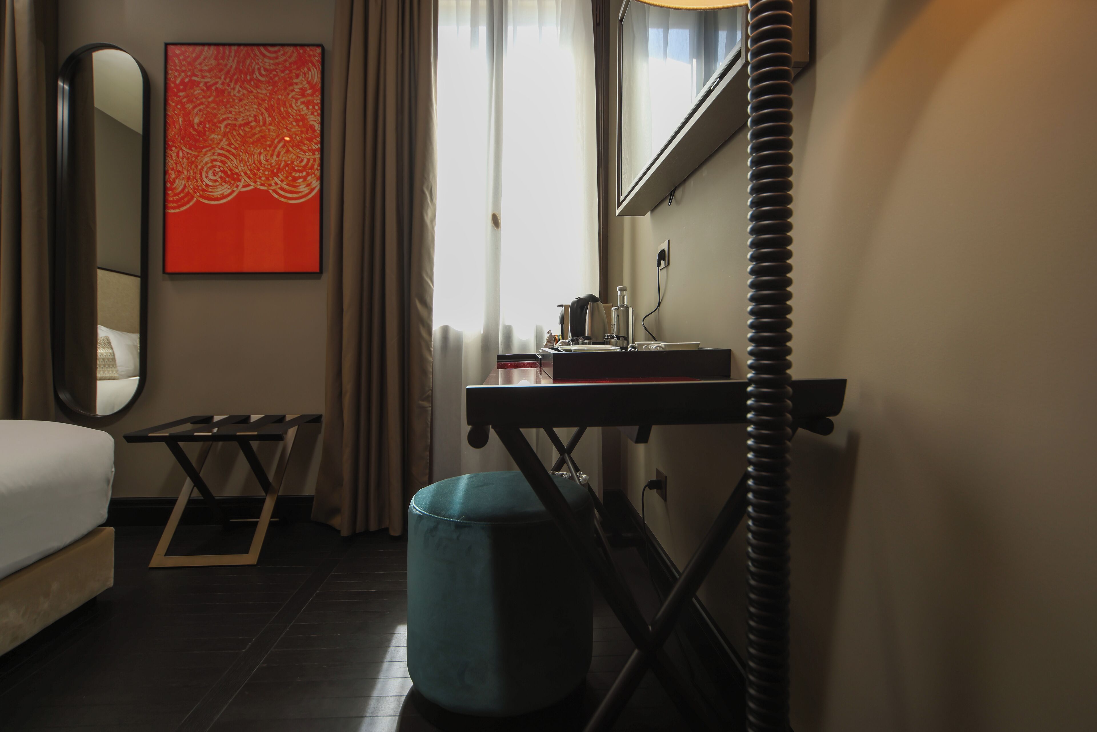 Une chambre d’hôtel avec un lit, un bureau, un miroir, une fenêtre avec des rideaux et un pouf de couleur turquoise.