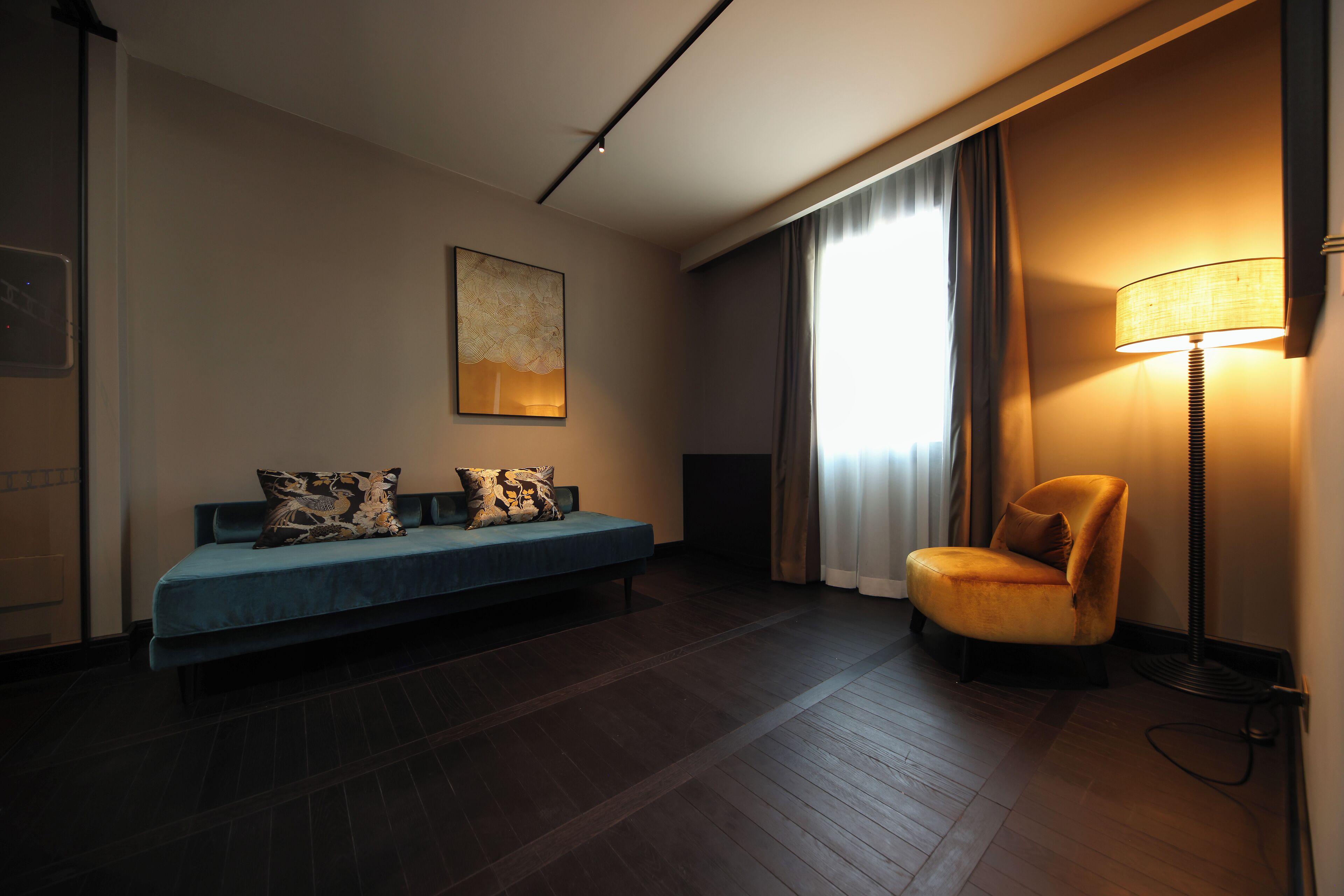 A hotel room with a dark wood floor, a blue sofa with patterned cushions, a single armchair, a standing lamp, and a large window with curtains.