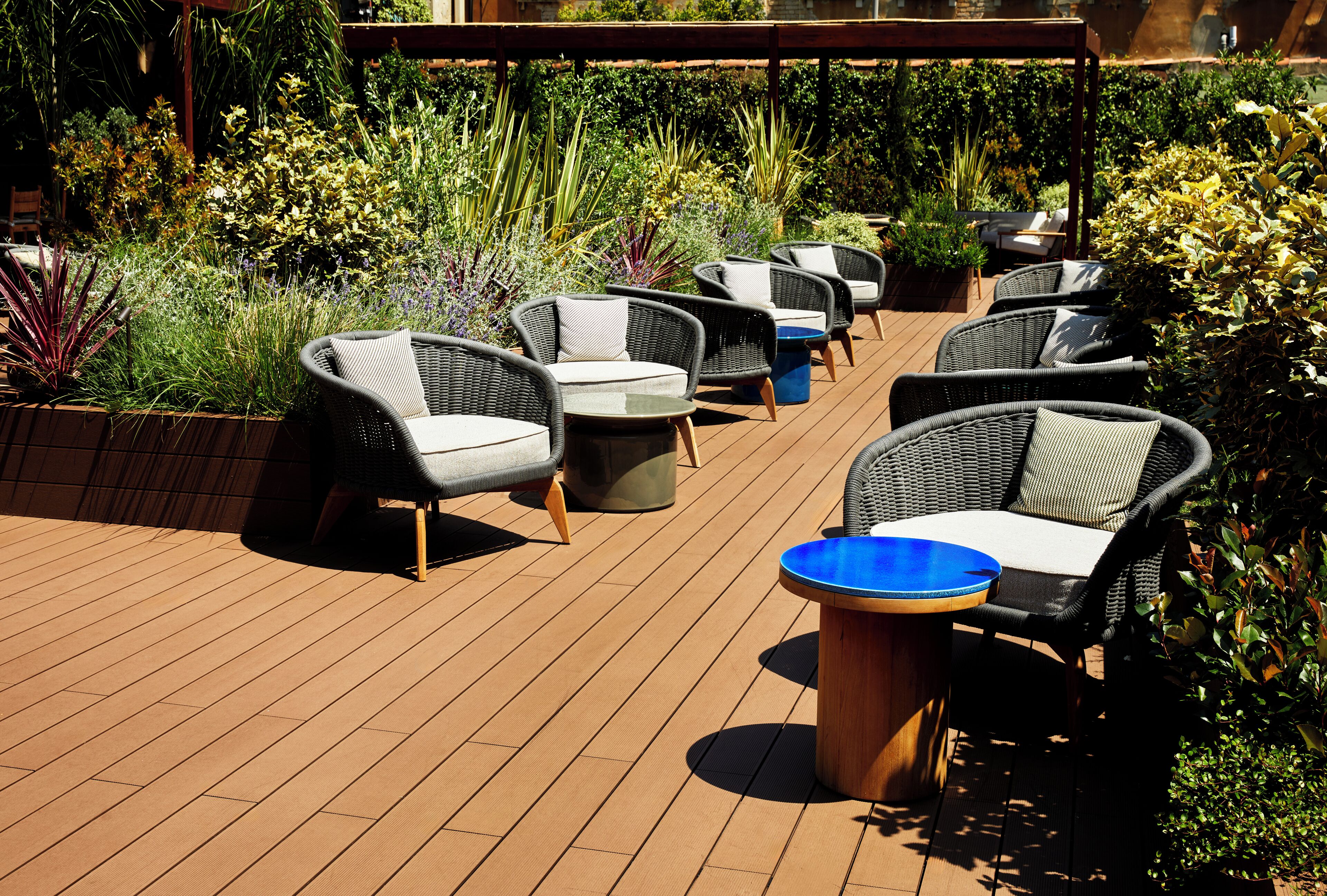 A deck with wicker furniture and a blue table, surrounded by various plants.
